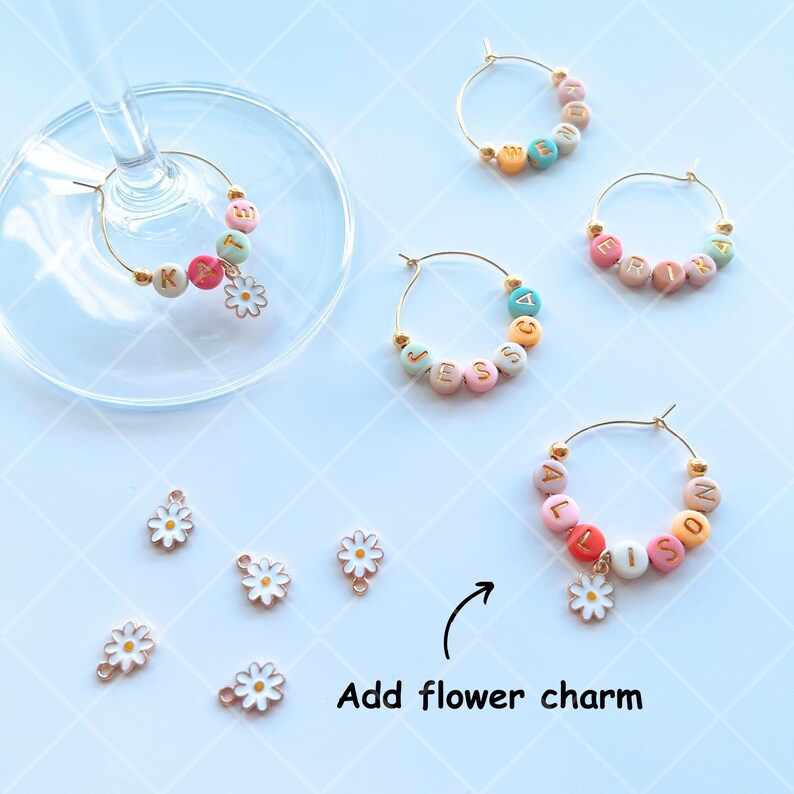 May include: Five gold-colored wine glass charms with colorful beads and letters spelling out names.  Each charm has a small white daisy charm attached.  The text "Add flower charm" is written below the charms.