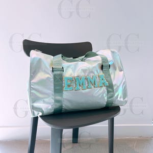 May include: A light blue iridescent duffel bag with a green zipper and straps. The bag has the name "EMMA" spelled out in turquoise letters with gold glitter.