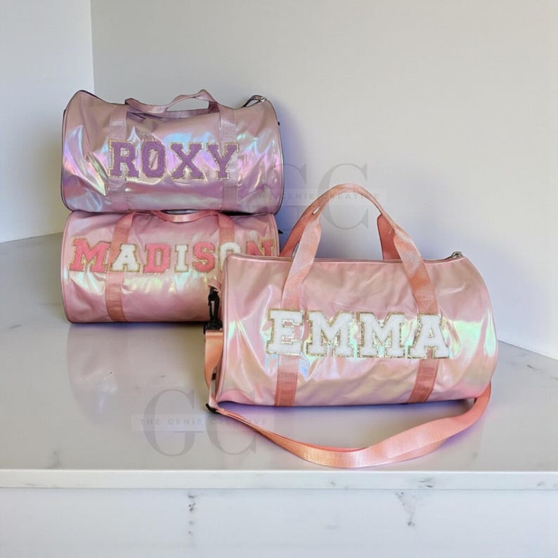 以下が含まれることがあります： ピンク色の光沢のあるダッフルバッグ3つ。バッグには「ROXY」、「MADISON」、「EMMA」の名前が白とピンクの刺繍文字でパーソナライズされています。各バッグにはハンドルとショルダーストラップが付いています。