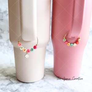 May include: Two stainless steel tumblers with personalized charm rings. The tumbler on the left is light beige with a charm ring that spells "TAYLOR" in colorful beads with a white daisy charm. The tumbler on the right is pink with a charm ring that spells "JESSICA" in colorful beads.