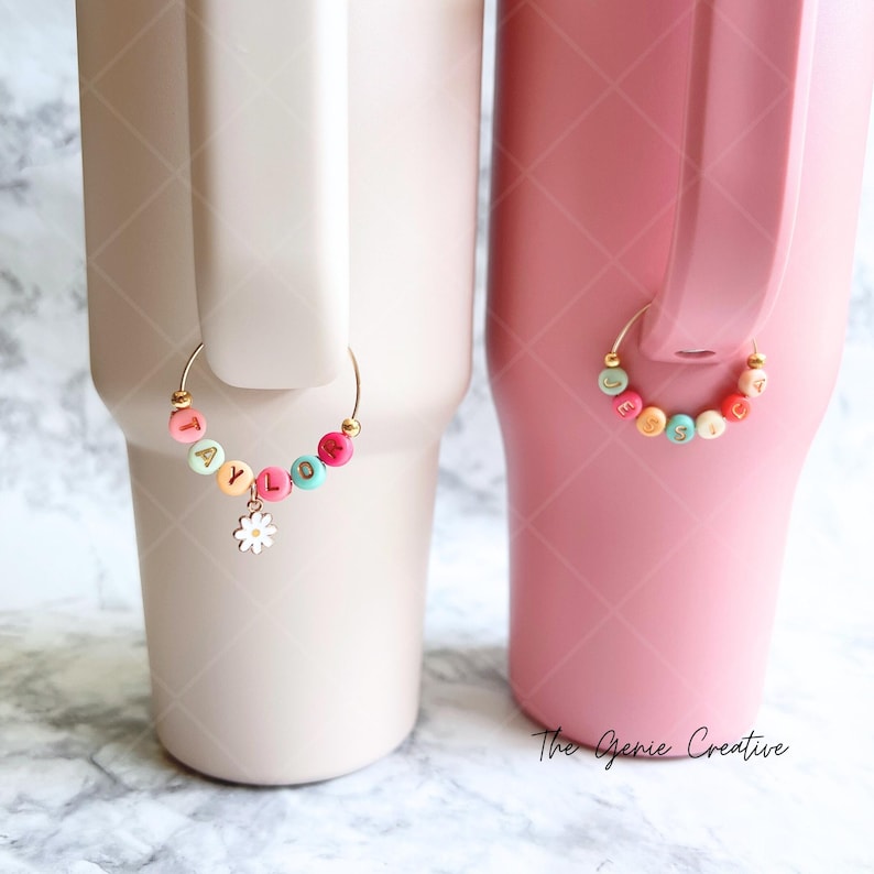 May include: Two stainless steel tumblers with personalized charm rings. The tumbler on the left is light beige with a charm ring that spells "TAYLOR" in colorful beads with a white daisy charm. The tumbler on the right is pink with a charm ring that spells "JESSICA" in colorful beads.