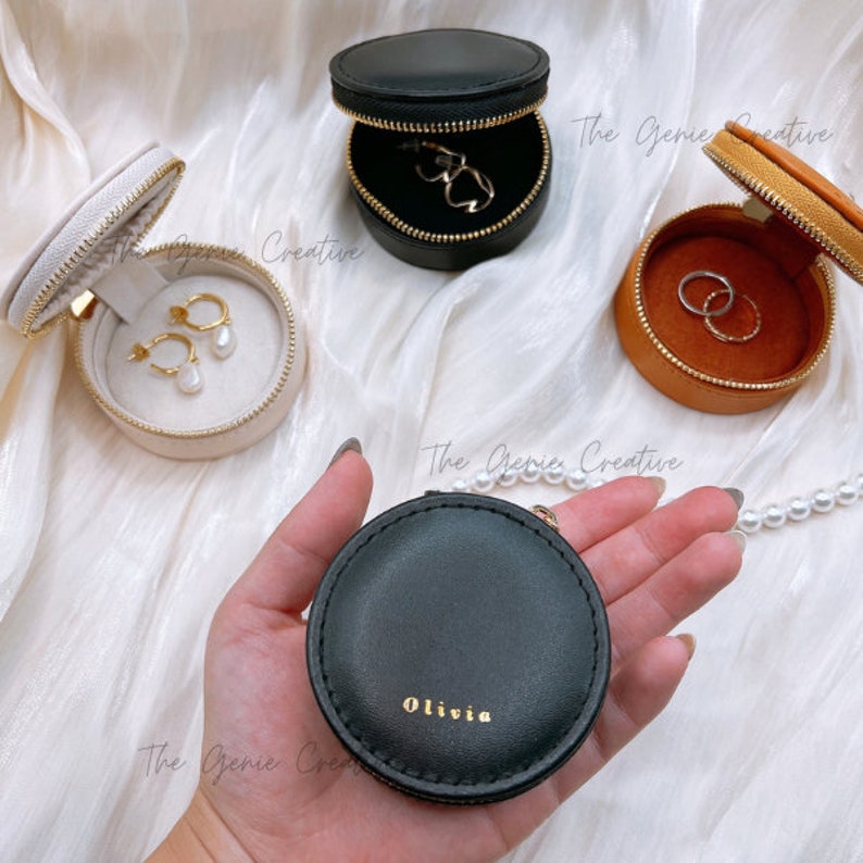 May include: Three round zippered jewelry boxes in black, beige, and brown. The black box has the word "Olivia" inscribed on the lid. The beige box contains a pair of pearl earrings. The brown box contains two gold rings.