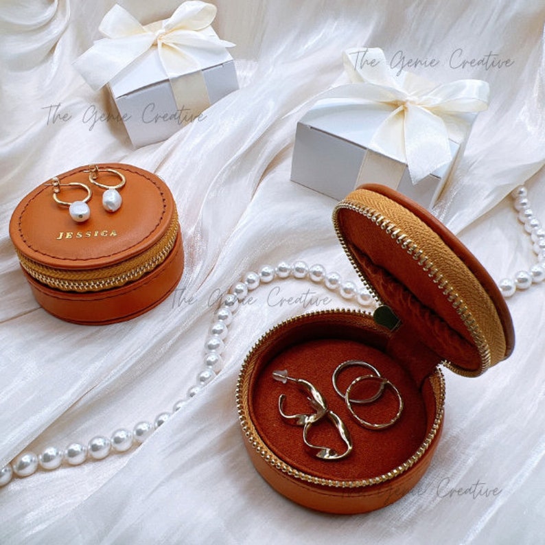 May include: Two round leather jewelry boxes with zippers. The smaller box is closed and has the name "JESSICA" engraved on the lid. The larger box is open and has a brown velvet interior. The box contains two pairs of gold earrings.