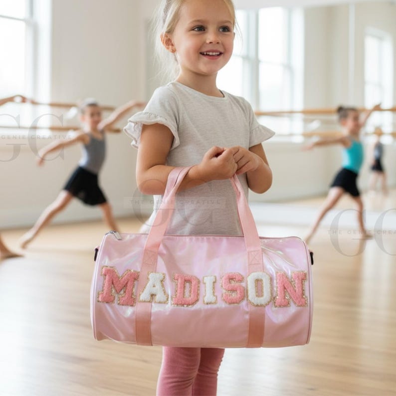 以下が含まれることがあります： 白とピンクの文字で「MADISON」と書かれたピンク色のダッフルバッグ。バッグにはピンク色のハンドルとトリムが付いています。画像には、ダンススタジオでバッグを持っている若い女の子が写っており、背景には他の子供たちがいます。