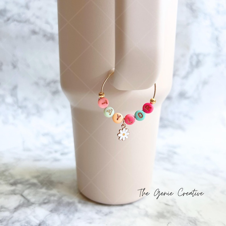 May include: A gold-colored charm with the name "TAYLOR" spelled out in colorful beads. The charm is attached to a gold-colored ring and has a small white daisy charm hanging from it.