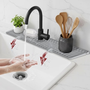 May include: A modern kitchen sink with a black faucet, gray drainboard, and white soap dish. Wooden kitchen utensils are in a dark gray ceramic container. A small potted plant adds a touch of green. Water flows from the faucet.