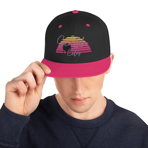 May include: Black baseball cap with a pink brim and a colorful embroidered design. The design features the text "Creatide City" above a sunset graphic.