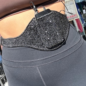 May include: A black sparkly belt with a black sparkly face mask attached to it. The belt is worn around the waist of a person wearing black leggings.