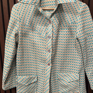May include: A vintage teal and tan checkered jacket with a wide collar, long sleeves, and two front pockets. The jacket has three buttons down the front and is hanging on a wooden hanger.
