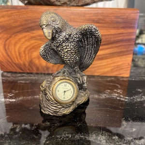 Könnte beinhalten: Bronze-farbene Papageienfigur mit ausgebreiteten Flügeln, die auf einer Felsformation sitzt. Der Sockel der Figur hat eine kleine Uhr mit weißem Zifferblatt und schwarzen Ziffern. Die Zeiger zeigen auf 12:00 Uhr.