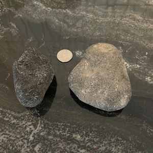 May include: Two smooth, gray rocks of different sizes on a black and white speckled surface. A silver coin is in the background.