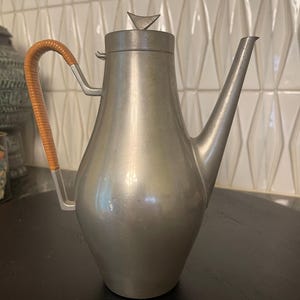 Pode incluir: Uma cafeteira vintage de metal prateado com um bico longo e uma alça de rattan trançado. A tampa apresenta um remate triangular. A cafeteira é exibida sobre uma superfície escura, mostrando seu design clássico. Uma peça decorativa para a cozinha.