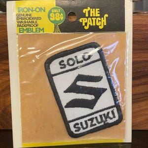 May include: An iron-on patch featuring the Suzuki logo in black and white, with the words "SOLO" and "SUZUKI" on a white background. The patch is packaged with the text "IRON-ON EMBLEM" and "The Patch".