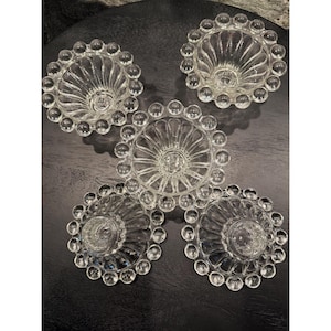 May include: A set of five clear glass bowls featuring a scalloped interior design and a decorative border of raised glass spheres around the outer rim. Each bowl has a circular base and a transparent finish, arranged in a cluster on a dark surface.