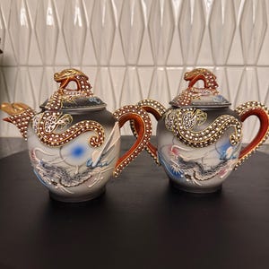 Op de afbeelding: Twee sierlijke theepotten met drakenmotieven. De theepotten hebben gouden accenten, rode handvatten en gedetailleerde drakenontwerpen op de deksels en lichamen. De achtergrond is een witte, getextureerde muur.