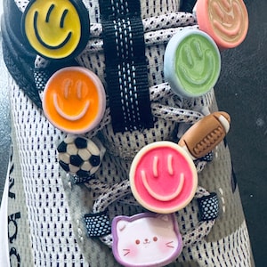 Puede incluir: Primer plano de una zapatilla deportiva con varios dijes coloridos para zapatos. Los dijes incluyen caras sonrientes, un balón de fútbol, un balón de fútbol americano y una cara de gato. La zapatilla tiene un diseño de malla gris y blanca con cordones blancos y negros.