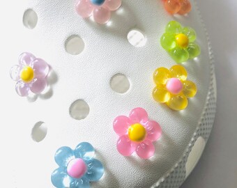 May include: A close-up of a white shoe with colorful flower-shaped charms. The charms are made of plastic and have a translucent finish. The charms are in various colors, including pink, blue, yellow, and green.