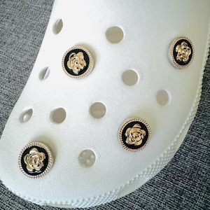 May include: Four gold-colored flower charms with black centers are attached to a white shoe. The charms are decorative and can be used to personalize shoes.