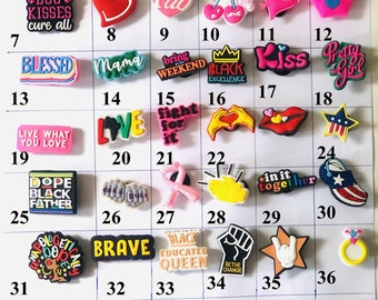 May include: A collection of colorful shoe charms with various designs and text. The charms include phrases like "Women Support Women," "Love," "Kiss," and "Brave." Other charms feature hearts, stars, and other symbols.