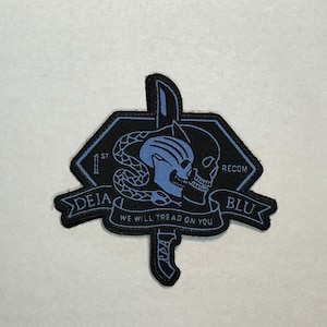 Może przedstawiać: Naszywka haftowana w kolorze czarnym i niebieskim z motywem czaszki i węża. Naszywka zawiera tekst "1st RECON DEJA BLU" i słowa "WE WILL TREAD ON YOU".
