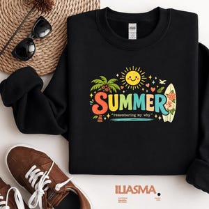 Könnte beinhalten: Schwarzes Sweatshirt mit einem farbenfrohen "SUMMER"-Aufdruck mit einer lächelnden Sonne, Palmen und einem Surfbrett. Der Text "remembering my why" steht unter dem Wort "SUMMER". Sonnenbrille und braune Turnschuhe sind im Hintergrund.