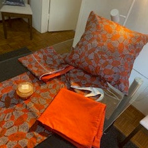 Op de afbeelding: Een oranje tafelkleed met wit patroon, twee bijpassende servetten en een kussen met hetzelfde patroon.