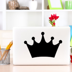 May include: A white laptop computer with a black crown sticker on the lid. The laptop is sitting on a wooden table with a pencil holder, a potted plant, and a book.
