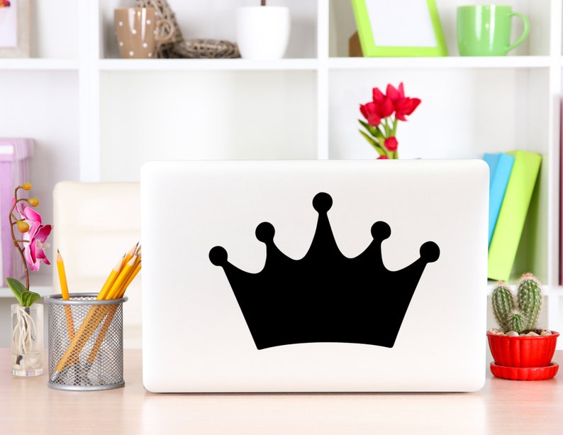 May include: A white laptop computer with a black crown sticker on the lid. The laptop is sitting on a wooden table with a pencil holder, a potted plant, and a book.