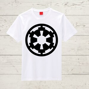 May include: White t-shirt featuring a large black Imperial symbol. The symbol is a circular design with a central circle and radiating shapes. The shirt has short sleeves and a crew neck, suitable for casual wear.