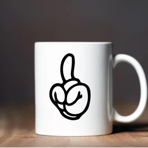 May include: White ceramic mug featuring a black cartoon hand gesture. The hand is making a rude gesture with the middle finger extended. The mug has a curved handle and is set against a blurred background.