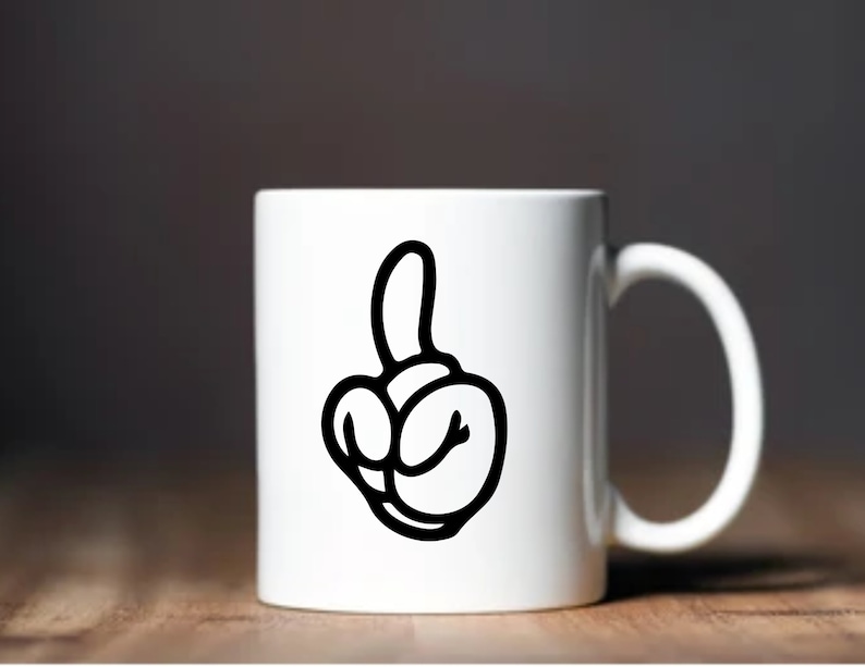 May include: White ceramic mug featuring a black cartoon hand gesture. The hand is making a rude gesture with the middle finger extended. The mug has a curved handle and is set against a blurred background.