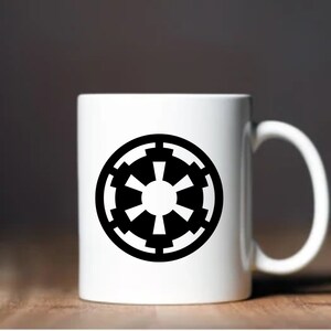May include: White ceramic mug featuring a black Imperial symbol. The mug has a curved handle and is set against a blurred background. The design is a bold, circular emblem, perfect for fans of science fiction.