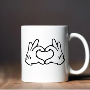 May include: White ceramic mug featuring a black and white graphic of cartoon hands forming a heart shape. The mug has a curved handle and is set against a blurred background.