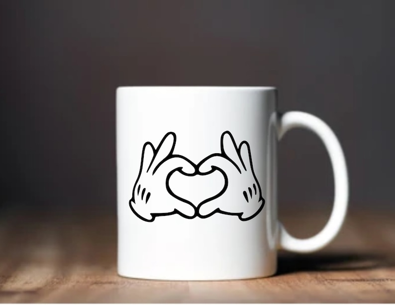 May include: White ceramic mug featuring a black and white graphic of cartoon hands forming a heart shape. The mug has a curved handle and is set against a blurred background.