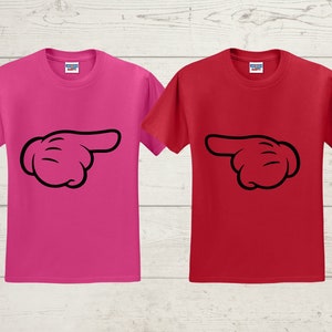 May include: Two short-sleeved t-shirts, one in bright pink and the other in red. Each shirt features a black cartoon hand pointing to the right. The shirts are displayed on a light wood-grain background.