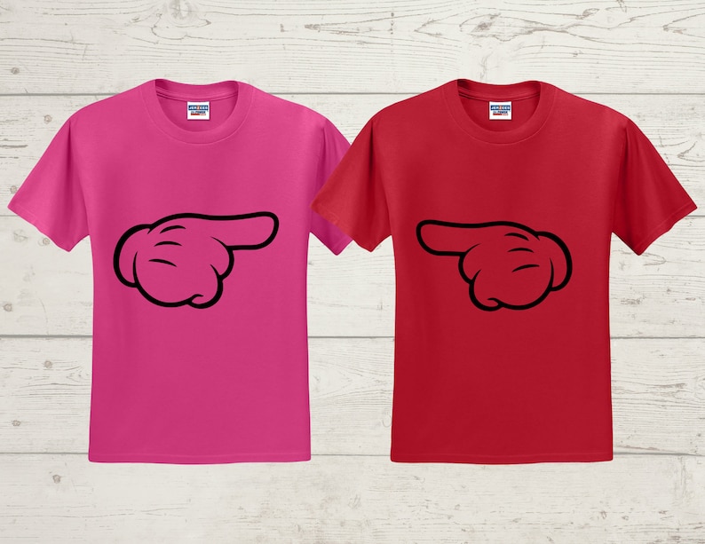 May include: Two short-sleeved t-shirts, one in bright pink and the other in red. Each shirt features a black cartoon hand pointing to the right. The shirts are displayed on a light wood-grain background.