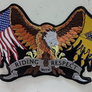 May include: A black embroidered patch with a bald eagle with outstretched wings, a tattered American flag, and a yellow triangle with the text "Patriot Guard Riders". The eagle is holding a banner with the text "Riding with Respect".