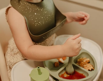Puede incluir: Un niño con un babero de silicona verde con estrellas blancas está sentado en una silla alta y come de un plato de silicona verde dividido con una cuchara de silicona verde. El plato tiene tres secciones, una con nuggets de pollo, otra con patatas fritas y otra con salsa roja. Un vaso de silicona verde con una pajita y un diseño de elefante está sobre la mesa.