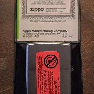 K&ouml;nnte beinhalten: Ein Zippo-Taschenfeuerzeug in einer schwarzen Schachtel. Das graue Feuerzeug hat ein rotes Warnetikett. Die Schachtel enth&auml;lt eine Garantiekarte mit Text. Das Zippo-Logo ist sichtbar.
