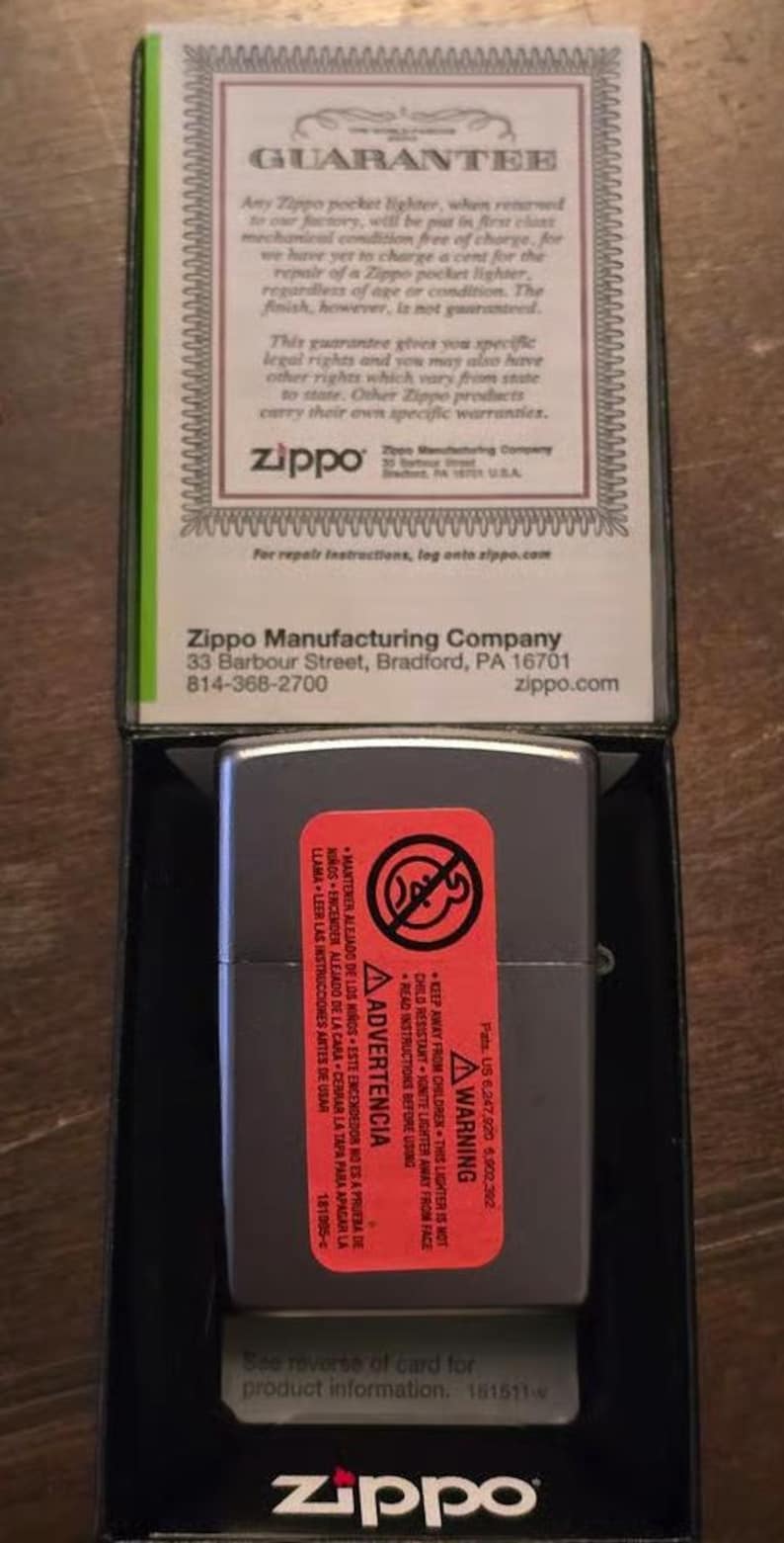 K&ouml;nnte beinhalten: Ein Zippo-Taschenfeuerzeug in einer schwarzen Schachtel. Das graue Feuerzeug hat ein rotes Warnetikett. Die Schachtel enth&auml;lt eine Garantiekarte mit Text. Das Zippo-Logo ist sichtbar.