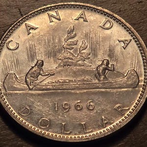 May include: A silver Canadian dollar coin from 1966. The coin features a canoe with two figures paddling, a stylized tree, and the word "CANADA" at the top. The word "DOLLAR" is at the bottom.