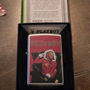 K&ouml;nnte beinhalten: Ein silbernes Zippo-Feuerzeug mit einem Playboy-Magazin-Cover-Design. Das Cover zeigt eine Frau in roter Kleidung. Das Feuerzeug befindet sich in einer schwarzen Box mit einer Garantiekarte.