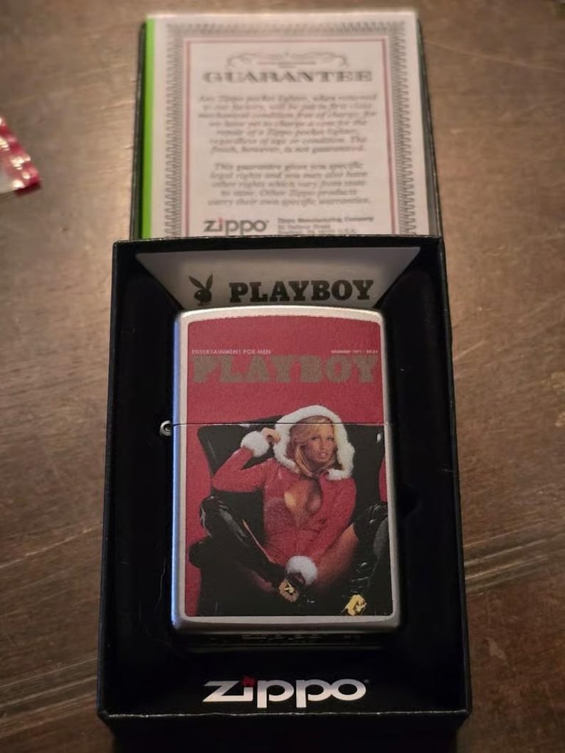 K&ouml;nnte beinhalten: Ein silbernes Zippo-Feuerzeug mit einem Playboy-Magazin-Cover-Design. Das Cover zeigt eine Frau in roter Kleidung. Das Feuerzeug befindet sich in einer schwarzen Box mit einer Garantiekarte.