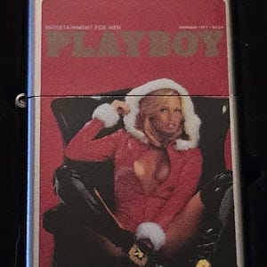 K&ouml;nnte beinhalten: Ein silbernes Metallfeuerzeug mit roter Abdeckung, die ein Vintage-Playboy-Magazin-Cover zeigt. Das Cover zeigt eine Frau in roter Kleidung mit einer Kapuze mit wei&szlig;em Pelzbesatz. Der Text "PLAYBOY" und "ENTERTAINMENT FOR MEN" sind sichtbar.