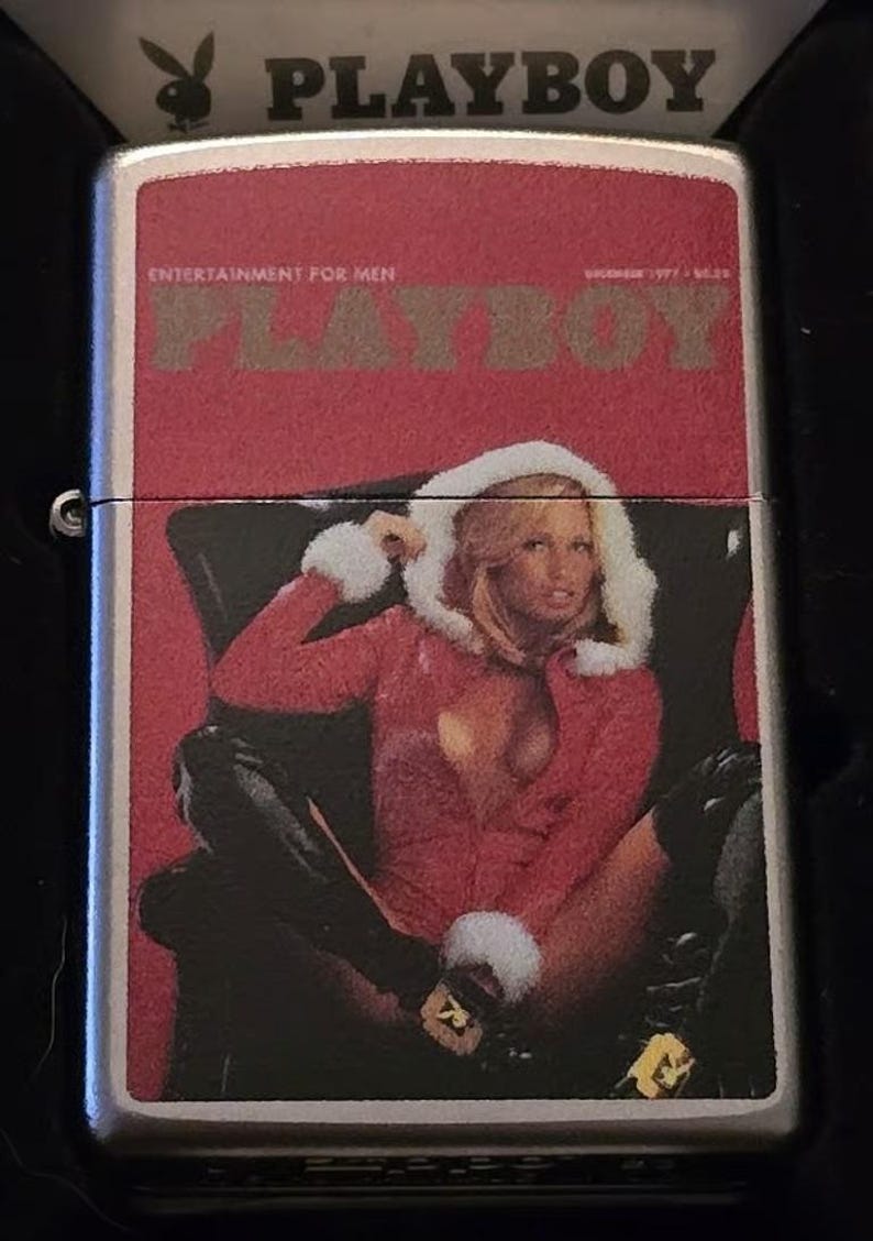 K&ouml;nnte beinhalten: Ein silbernes Metallfeuerzeug mit roter Abdeckung, die ein Vintage-Playboy-Magazin-Cover zeigt. Das Cover zeigt eine Frau in roter Kleidung mit einer Kapuze mit wei&szlig;em Pelzbesatz. Der Text "PLAYBOY" und "ENTERTAINMENT FOR MEN" sind sichtbar.