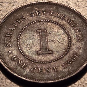 May include: A close-up of a worn, circular coin. The coin features the text "STRAITS SETTLEMENTS" around the top edge, "ONE CENT" at the bottom, and the year "1886". A large "1" is in the centre.