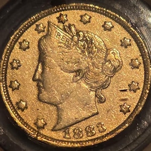 May include: A gold coin featuring a profile of a woman with the word "LIBERTY" above her head. The coin is dated 1883 and has a ring of stars around the edge. The coin is in a protective case.