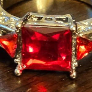 May include: A silver ring with a large, square, red gemstone surrounded by smaller, triangular, red gemstones. The ring band is decorated with a silver filigree design.