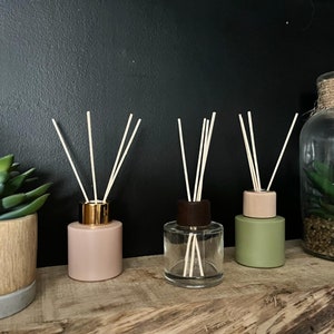 May include: Three glass diffuser bottles with wooden tops and natural reed diffusers. The bottles are filled with a pink, clear, and green liquid. The bottles are sitting on a wooden surface.