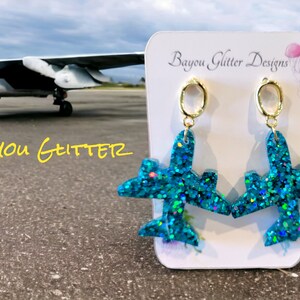 May include: A pair of turquoise blue glitter aeroplane earrings with gold hoops. The earrings are on a white card with the text "Bayou Glitter Designs".